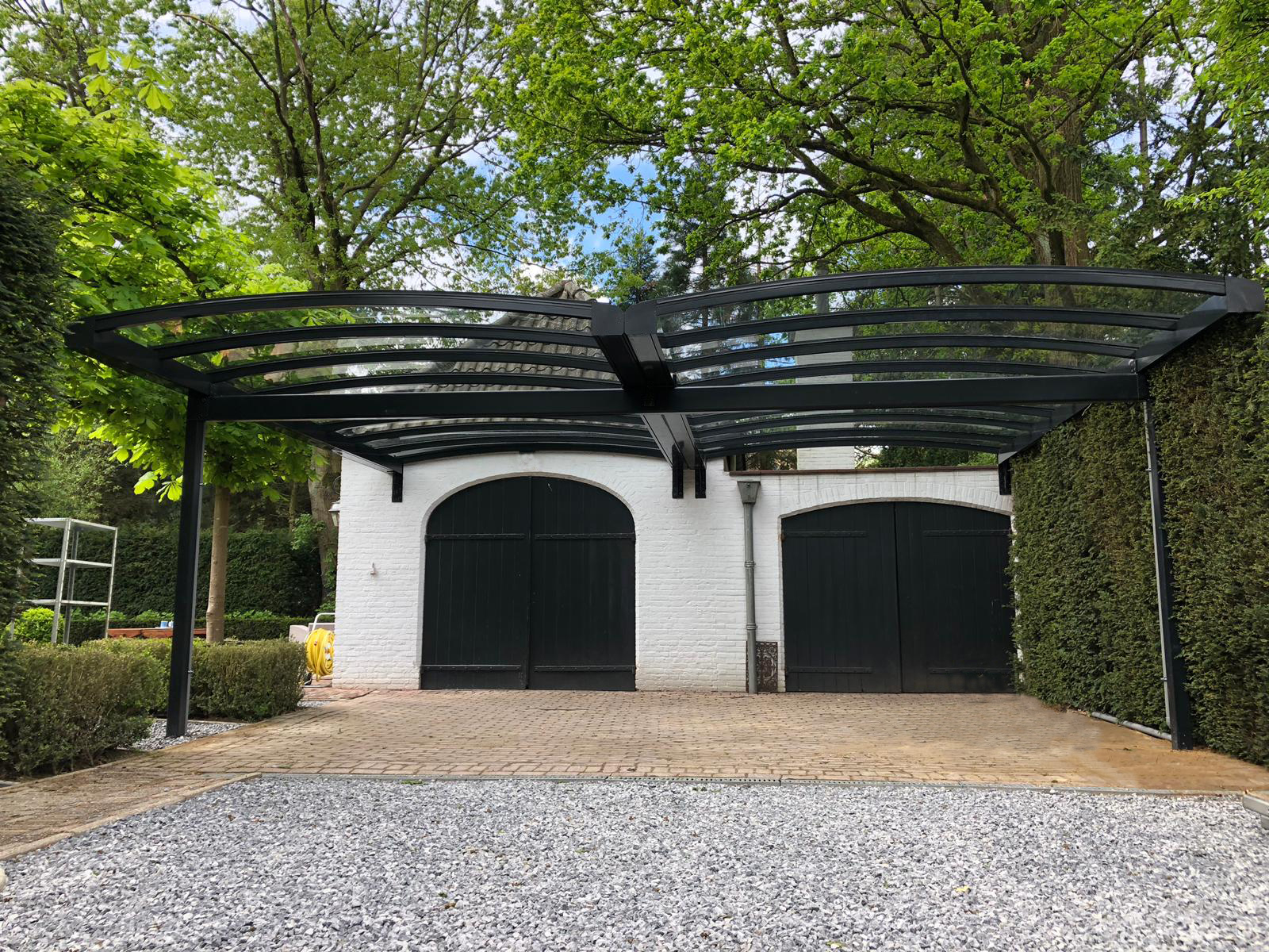 Dubbele gebogen aluminium carport met glas laten plaatsen tegen witte stenen garage met groene deuren.