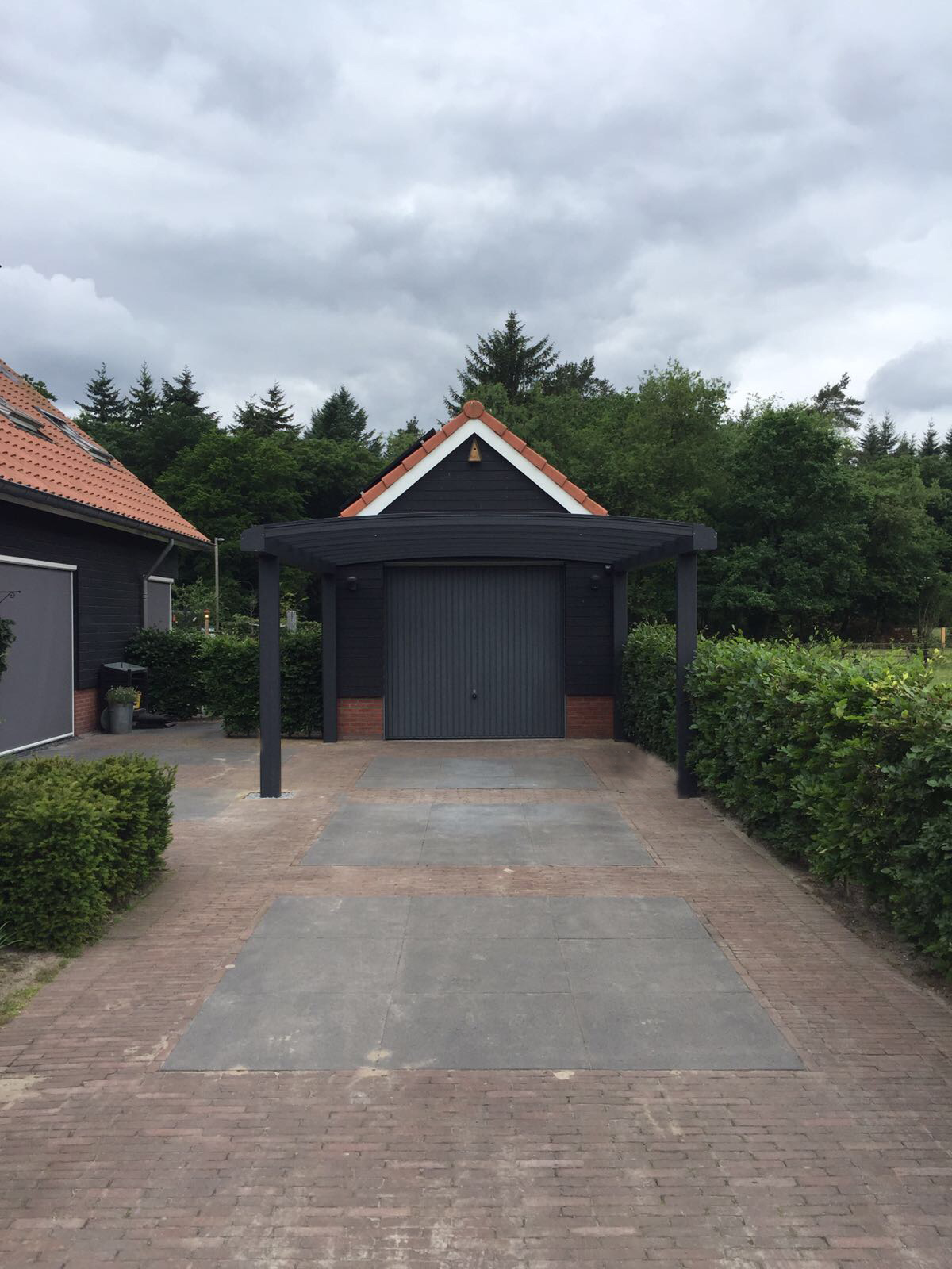 Vooraanzicht van grijze gebogen carport tegen zwarte houten garage