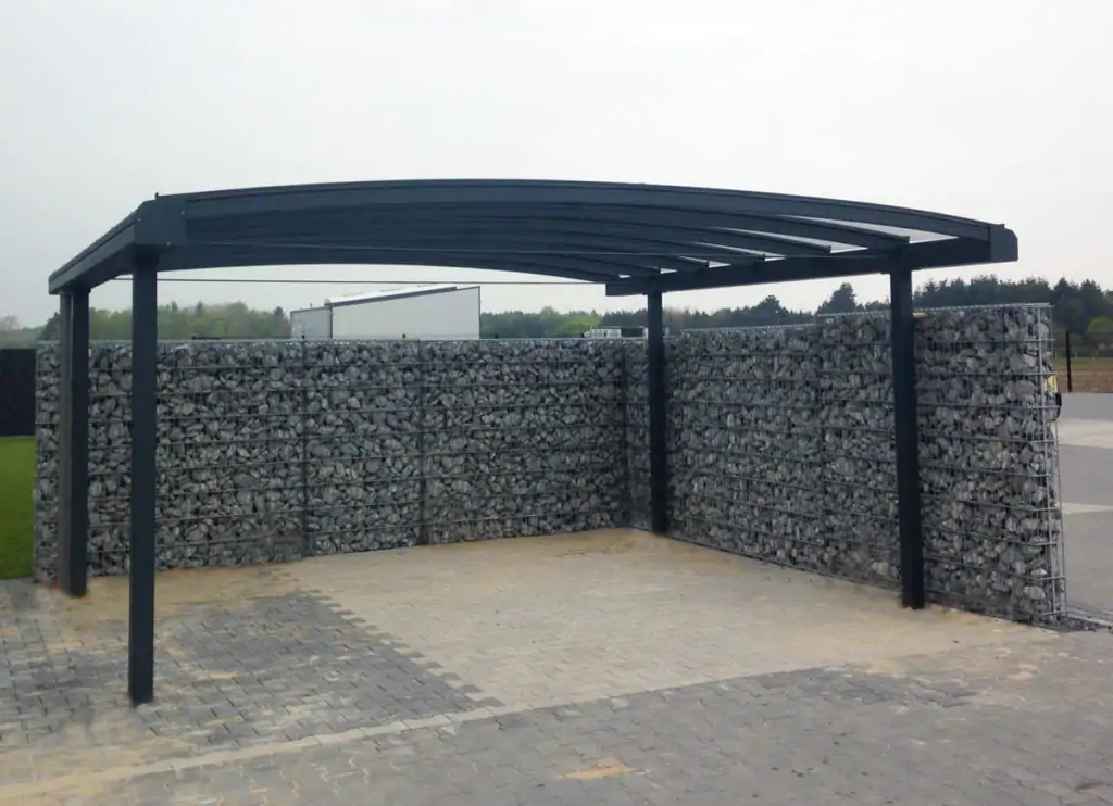 Gebogen type prefix aluminium carport overkapping met glas. Omheind met schanskorfmuur met grijze stenen.
