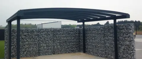 Aluminium carport met gebogen dak en glazen overkapping, omringd door een schanskorfmuur gevuld met grijze stenen.